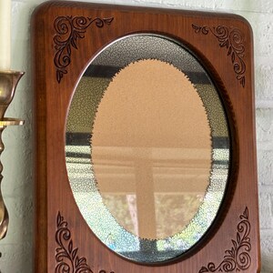 May include: A rectangular wooden mirror frame with rounded corners and an oval center opening. The wood features carved scrollwork patterns in each of the four corners. The mirror glass has a distinct crackled, aged texture around the perimeter.