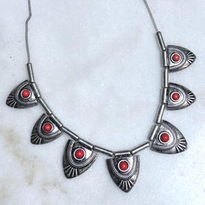 May include: A silver-toned necklace featuring six triangular metal pendants, each inlaid with a small, round, vibrant red stone. The pendants are connected by smooth silver-toned cylindrical beads and suspended from a thin silver-toned chain.