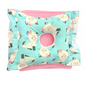 May include: A turquoise pillow with a white cat pattern and pink trim. The pillow has a hole in the center for a nursing mother's breast.
