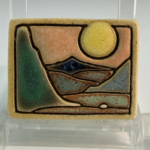 May include: A square ceramic tile featuring a landscape scene with a yellow sun, mountains, and water. The tile has a beige border and is supported by a clear plastic stand. The landscape includes green, blue, and brown tones.