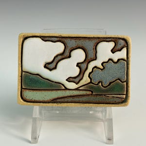 May include: Rectangular ceramic tile with a landscape design. The tile features a sky with white cloud shapes, a gray-blue background, and green land. The tile has a tan border and is displayed on a clear stand.