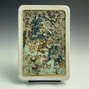 May include: Rectangular ceramic art piece with a white border and a textured, abstract design. The artwork features a mix of colors, including blue, green, brown, and gold, creating a unique visual texture. The piece is mounted on a black stand.