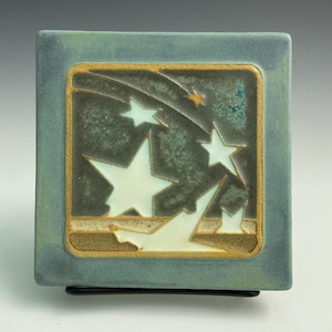 May include: A square ceramic tile with a blue-green glaze. The tile features a design of white stars and streaks of white against a dark green background. The tile is framed with a gold border.