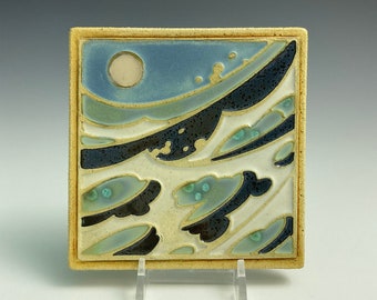 Könnte beinhalten: Eine Keramikfliese mit einem blauen und weißen Wellendesign. Die Fliese hat einen weißen Mond in der oberen linken Ecke und ein schwarz-weißes Wellendesign in der unteren rechten Ecke.