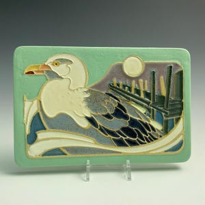 May include: A rectangular ceramic tile with a seagull design. The seagull is white and gray with a yellow eye and red beak, set against a light green background. The design includes a moon, industrial structures, and waves, with gold accents.