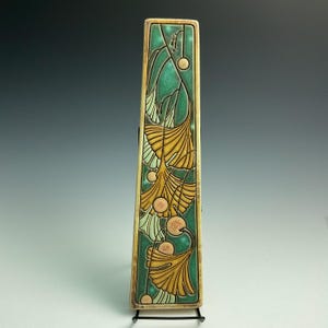 May include: A tall, rectangular ceramic tile featuring a relief design of golden ginkgo leaves against a deep teal background. The piece has a raised border and is displayed on a black metal stand, showcasing intricate botanical line work.