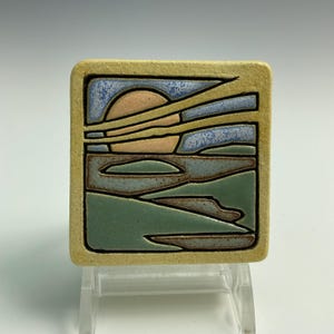 May include: A ceramic tile with a sunset scene. The tile is square and has a light yellow border. The sunset is depicted in shades of orange, yellow, and blue. The sky is a light blue with yellow clouds. The sun is a bright orange circle. The horizon is a dark blue line. The water is a light blue with a few waves. The sand is a light brown. The tile is a handmade art piece.