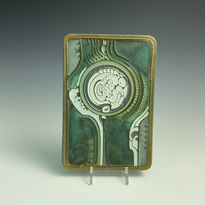 May include: Rectangular ceramic tile with a green and white abstract design. The tile has a bronze-colored border and features a central circular motif with organic shapes. The tile is displayed on a clear stand.