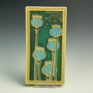 May include: A rectangular ceramic tile featuring a relief design of four light blue poppy seed pods on long stems. The background is a deep, textured forest green with a crackle glaze finish. The tile is framed by a raised, matte cream-colored border.