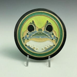 May include: A ceramic coaster with a green, blue, and white frog design. The frog has black eyes and a yellow mouth. The coaster is surrounded by a black border and a green and gold ring.