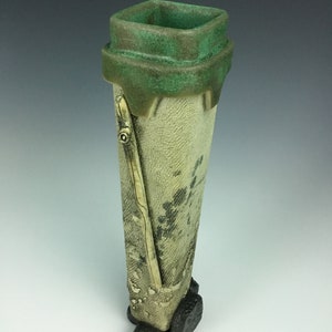May include: A tall, ceramic vase with a square opening and a green glaze. The vase has a textured, off-white body with black and brown accents. The base of the vase has four small, black feet.