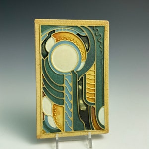 May include: A ceramic tile with an abstract design in shades of blue, green, yellow, and white. The tile features a central circle with a white interior and a blue border.