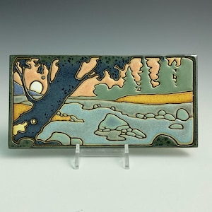 May include: A ceramic tile with a stylized landscape scene. The tile features a blue tree with a yellow sun in the background, a blue river with green and brown rocks, and a green and yellow hillside.