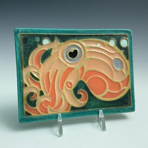 May include: A rectangular ceramic tile featuring a stylized octopus design in shades of peach and coral against a deep teal background. The octopus has a prominent eye with a heart-shaped pupil and swirling tentacles, all outlined in raised gold lines.