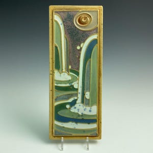 May include: Rectangular ceramic tile featuring a waterfall scene. The artwork showcases cascading water in shades of green and blue, set against a purple and tan background. A golden frame surrounds the image, and a sun-like design is at the top.