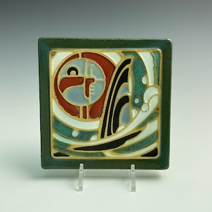 May include: Square ceramic tile with a green border and an abstract design. The design features red, black, white, and teal shapes with gold accents. The tile is displayed on a clear stand.