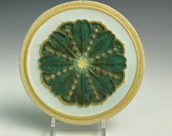 Könnte beinhalten: Ein runder Keramikuntersetzer mit einem grünen und goldenen Blumendesign. Der Untersetzer hat einen weißen Hintergrund und einen goldenen Rand.