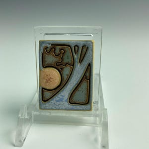 May include: A rectangular ceramic tile with a tree design. The tile features a light blue background with brown and tan accents. The tree design is outlined in brown, and a tan circle is on the left side.
