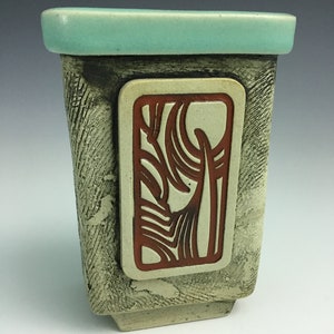 May include: A rectangular ceramic vase with a light green top and a textured, off-white body. A square, red-brown design is inset into the front of the vase.