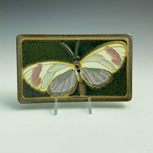 May include: A rectangular ceramic tile featuring a central butterfly motif. The butterfly has cream-colored wings with accents of soft pink and muted blue, set against a dark, speckled forest green background with a brown border and gold outlines.