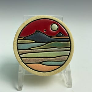 May include: A round ceramic tile featuring a landscape design. The image includes a red sky with a moon and a small dot, a dark blue mountain, and layered fields of green, tan, and blue. The tile has a beige border and black outlines.