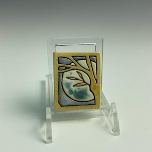 May include: A rectangular ceramic tile with a nature-themed design. The tile features a tree branch with leaves against a blue and white background, framed by a tan border. The tile is displayed on a clear plastic stand.