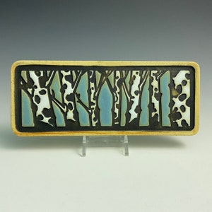 May include: A rectangular ceramic tile with a black border and a design of stylized trees in shades of blue and green.