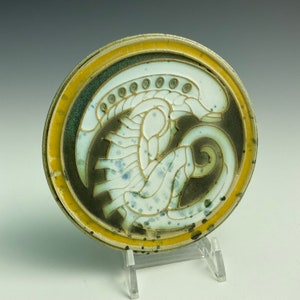 May include: A ceramic coaster with a white and green alien-like creature design on a dark green background. The coaster has a yellow rim.