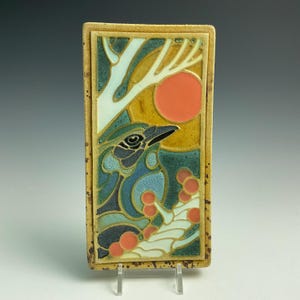 May include: A rectangular ceramic tile featuring a relief design of a blue bird perched among branches with red berries. The background includes a yellow sky and a large orange sun. The tile has a speckled tan border and a raised, textured surface.