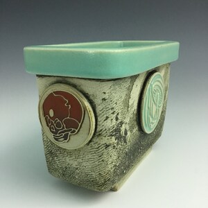 May include: A rectangular ceramic planter with a light blue glaze. The planter has three circular designs on the side, one with a red and white floral design, one with a green and white floral design, and one with a white floral design.