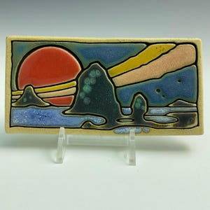 May include: Rectangular ceramic tile featuring a sunset over the ocean. The artwork depicts a large red sun, blue water, and dark green rock formations. The tile is framed with a beige border.