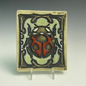 May include: A ceramic tile with a decorative design featuring a stylized ladybug in red, black, and white. The tile is framed with a black and gold design.
