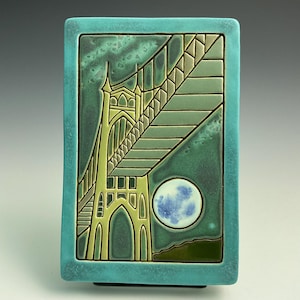 May include: A rectangular ceramic tile featuring a bridge design in shades of green and yellow against a teal background. The bridge is depicted with intricate details, and a full moon is visible in the lower right corner. The tile is framed by a teal border.