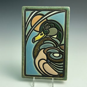 May include: A ceramic tile with a stylized image of a duck in shades of green, brown, and pink. The duck is surrounded by a blue and green background with a stylized sun.