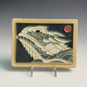 May include: A ceramic tile with a white and blue horned lizard design on a black background. A red circle is in the upper right corner of the tile.
