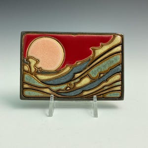 May include: A rectangular ceramic tile featuring a glossy glaze design of a pale peach sun set against a deep red sky. Below the sun, stylized waves in shades of muted blue, cream, and sage green are separated by dark, raised outlines.