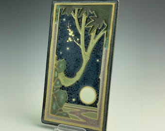 May include: A rectangular ceramic tile featuring a night landscape with a dark blue speckled sky, a pale yellow full moon, and a stylized tree with light green branches. The border is framed in dark gray with a thin yellow inner line.