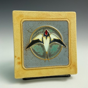 May include: A ceramic tile with a yellow frame and a blue background. The tile features a stylized white bird with black wings and a red beak, flying inside a circle with a green and blue background.