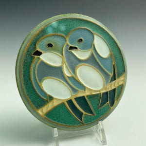 May include: A ceramic tile with a green background and two blue birds perched on a gold branch. The birds have white accents and are stylized in a geometric pattern.