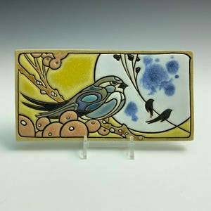 May include: A ceramic tile with a yellow, blue, and white background featuring a stylized bird perched on a branch with berries. The bird has blue, green, and brown markings. Two smaller black birds are flying in the background.