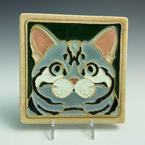 May include: A ceramic tile with a hand-painted image of a blue tabby cat with green eyes. The cat is looking directly at the viewer. The tile is surrounded by a beige border.