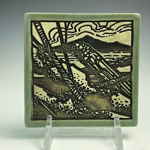 May include: A black and white ceramic tile with a stylized image of a landscape. The tile has a green border and is on a white stand.