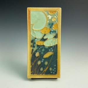 May include: A rectangular ceramic tile with a gold frame and a stylized floral design. The design features green, blue, and yellow colors.