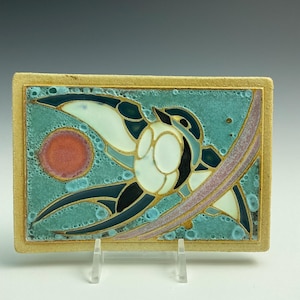 May include: Rectangular ceramic tile with a bird design. The bird is depicted with white, black, and teal details against a teal background. A red circle and a purple curved line are also present. The tile has a tan border and is displayed on a clear stand.