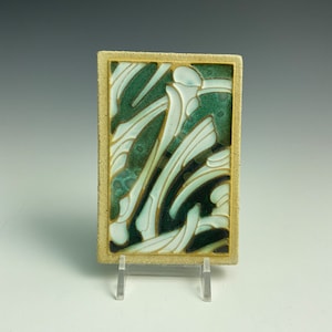 May include: A ceramic tile with a green, white, and black abstract design. The tile has a textured surface and a gold border.