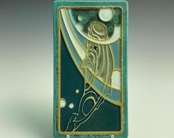 Könnte beinhalten: Rechteckige Keramikfliese mit türkisfarbenem Rand und stilisiertem Design. Das Kunstwerk zeigt eine zentrale Figur mit Hut, vor einem Hintergrund aus Türkis und Weiß, mit goldenen Akzenten. Das Design erinnert an den Jugendstil.
