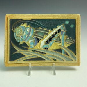 May include: A rectangular ceramic tile featuring a relief design of a grasshopper in shades of blue, yellow, and dark green. The insect is depicted amidst stylized blades of grass against a dark background, framed by a textured beige border.