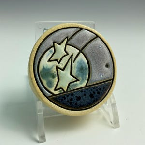 May include: A round ceramic pendant with a celestial design. The pendant features a cream-colored border, enclosing a design with two light green stars, a blue and green moon, and a gray and blue background. The bottom section is dark blue.