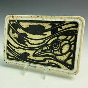 May include: A rectangular ceramic dish with a white background and black design. The design features a stylized bird with a large eye and a swirling pattern.