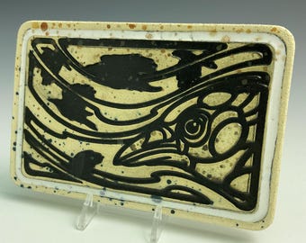 May include: A rectangular ceramic dish with a white background and black design. The design features a stylized bird with a large eye and a swirling pattern.